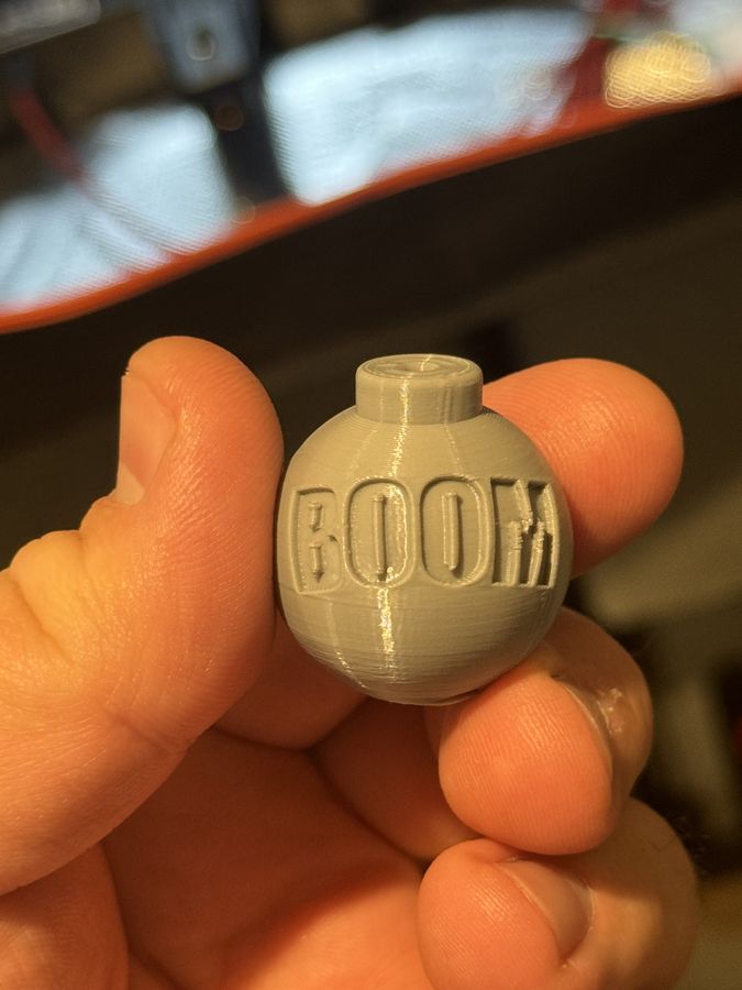 A tan 3D-printed round counterweight embossed with the word BOOM, held between two fingers