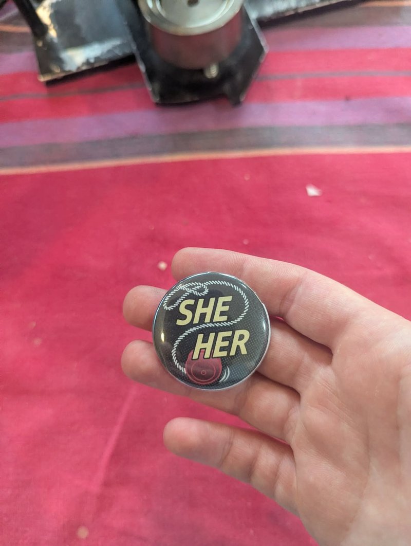 Pronoun pin reading SHE HER with a yo-yo design, one of a set