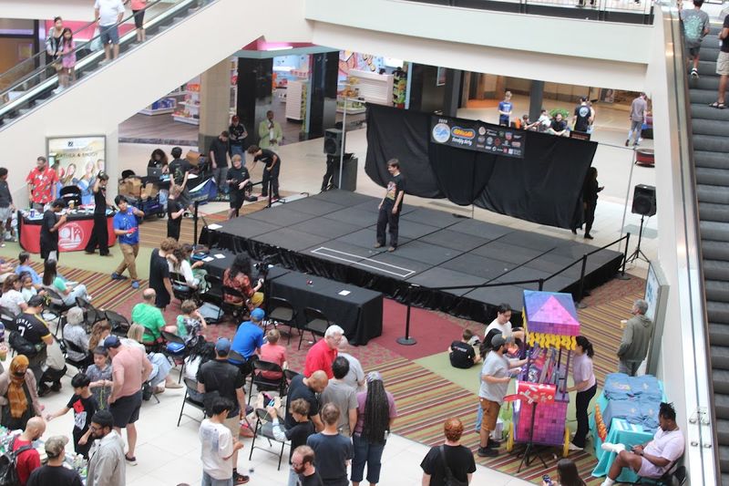Overhead view of Center Court at Dulles Town Center during the contest: a competitor on the black stage, judges at their table, and a crowd filling the floor and escalators