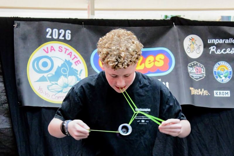 A competitor with curly hair holds the string in his teeth and stretches it into a triangle around the yo-yo in front of the VA State banner