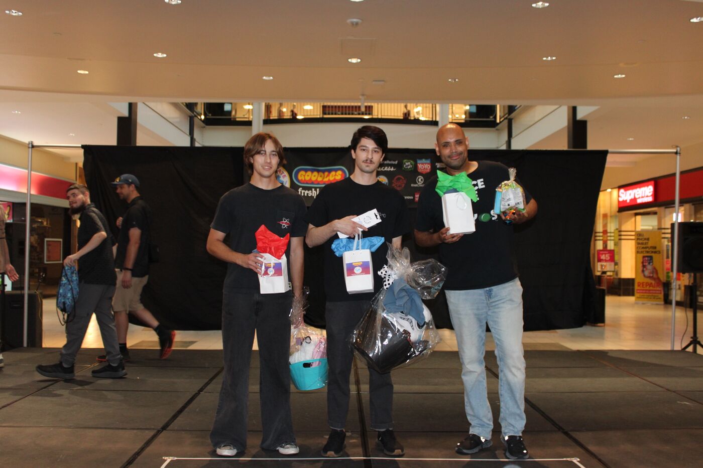X Division podium finishers Chandler Steele, Drew Buscemi and Richard Pigkaso stand on the Center Court stage holding prize bags and gift baskets, with the sponsor banner behind them