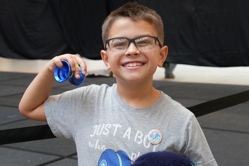 A young Sport, Beginner, and Junior competitor in glasses grins and holds up a blue yo-yo by the stage