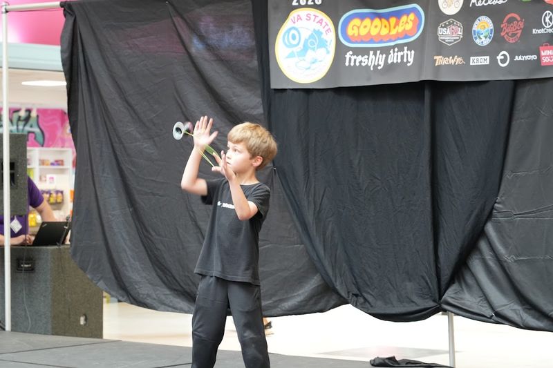 A young competitor on stage looks up at a yo-yo flying over his raised hands in front of the sponsor banner