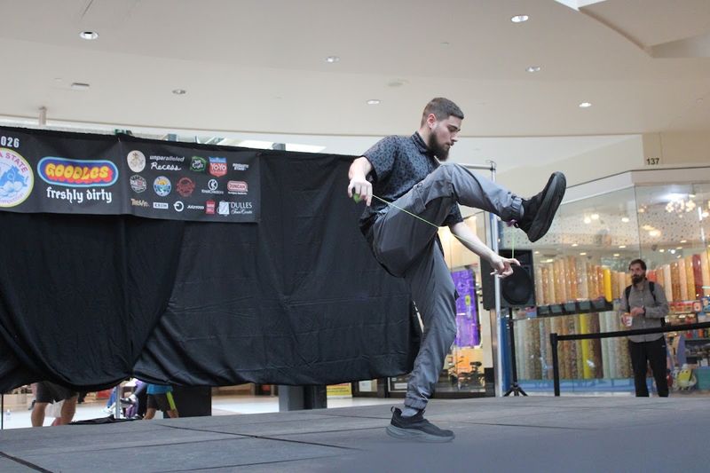 A competitor in gray kicks one leg high over the string mid-trick in front of the sponsor banner