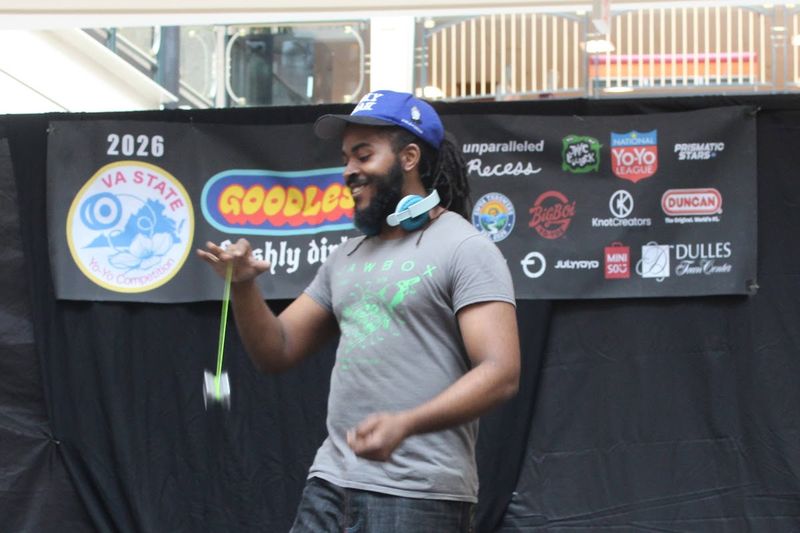 A competitor in a gray shirt and blue cap smiles while a yo-yo spins at his side in front of the VSYC-26 sponsor banner