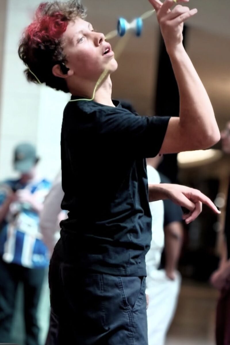 A competitor with red-tipped curly hair looks up at a blue yo-yo spinning at eye level during a trick