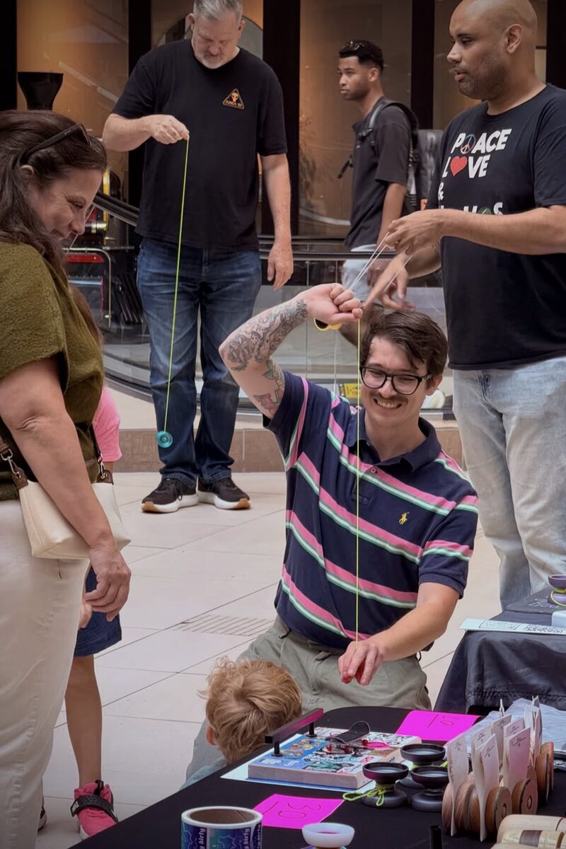 A thrower in a striped polo kneels at a vendor table to show a yo-yo to a family while two more throwers practice behind