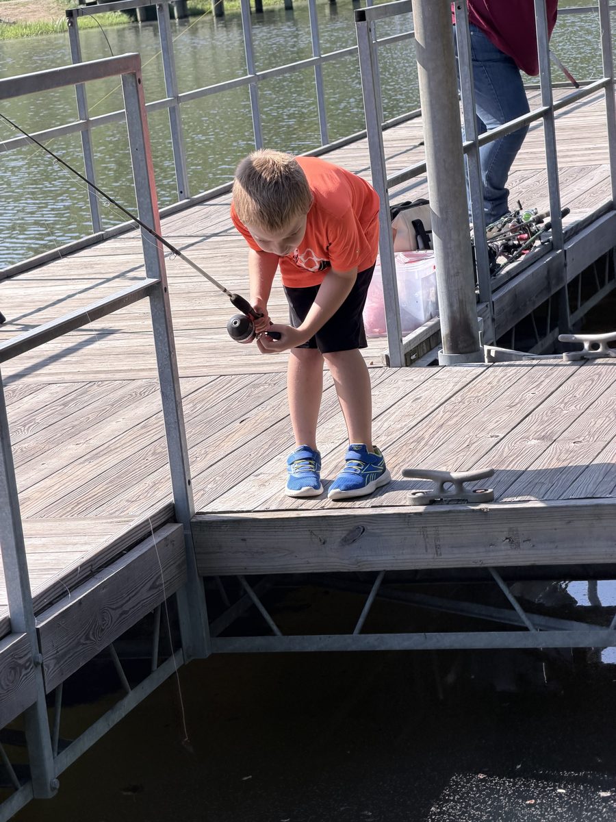 A Cub Scout in an orange shirt leans over a dock railing, reeling in a fishing line.