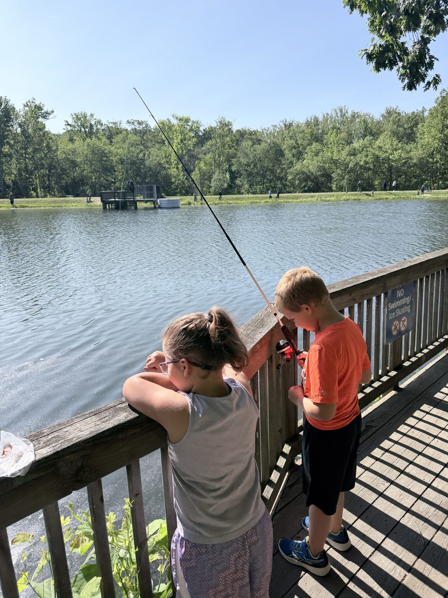 Two Cub Scouts fish from a wooden boardwalk over a sunny pond lined with trees.