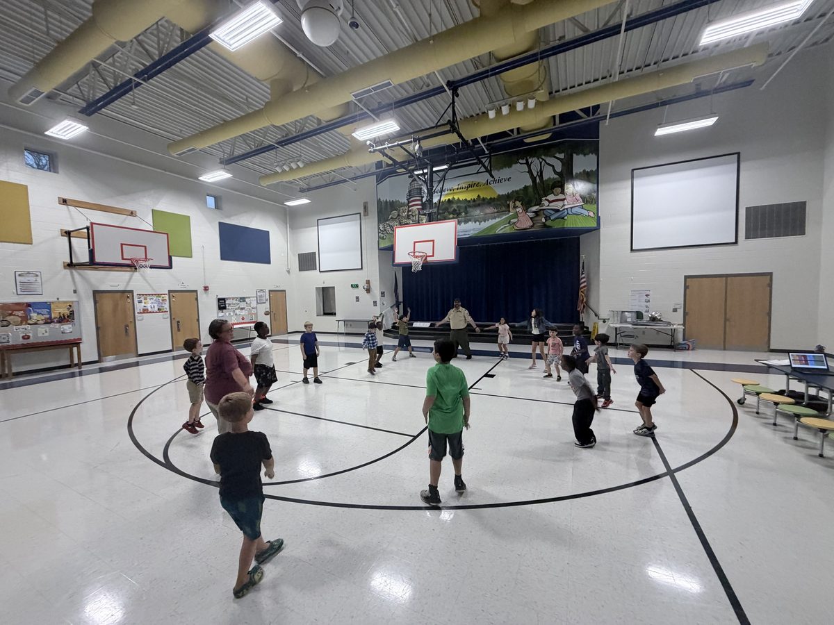 Cub Scouts spread out in a circle on the school gym floor with a uniformed leader and a parent, stretching and moving together.