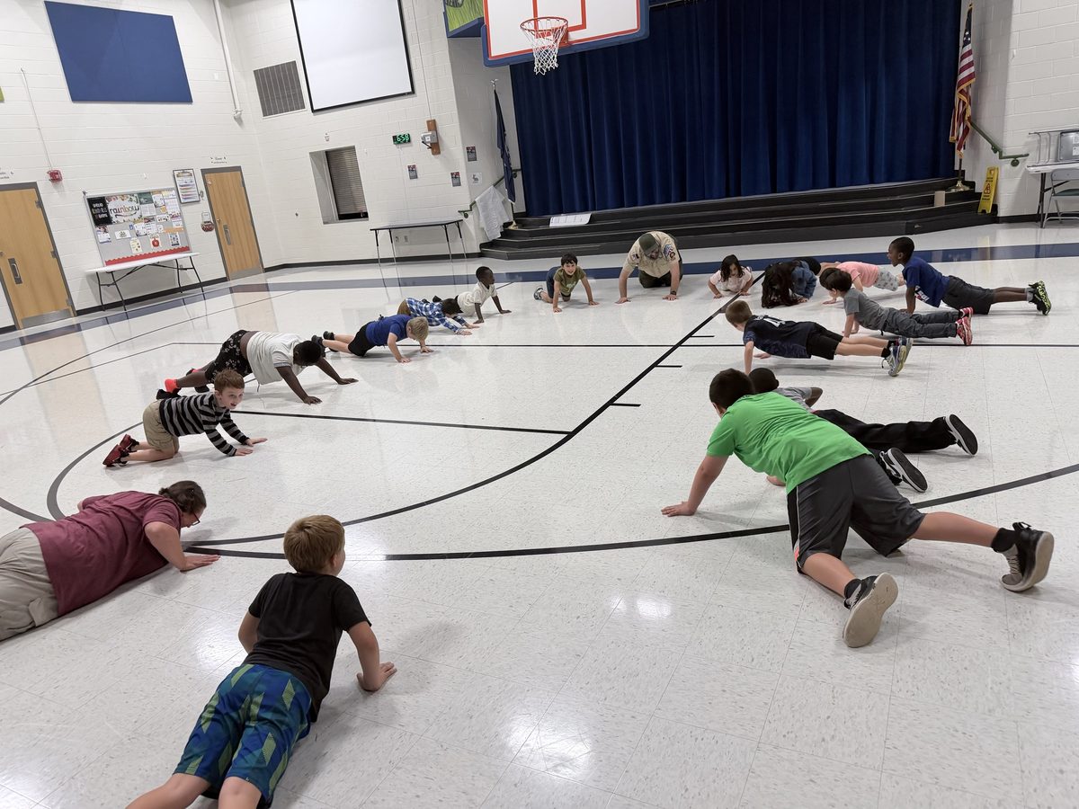 Scouts and leaders do push-ups and bear crawls in a big circle on the gym floor, smiling at each other.