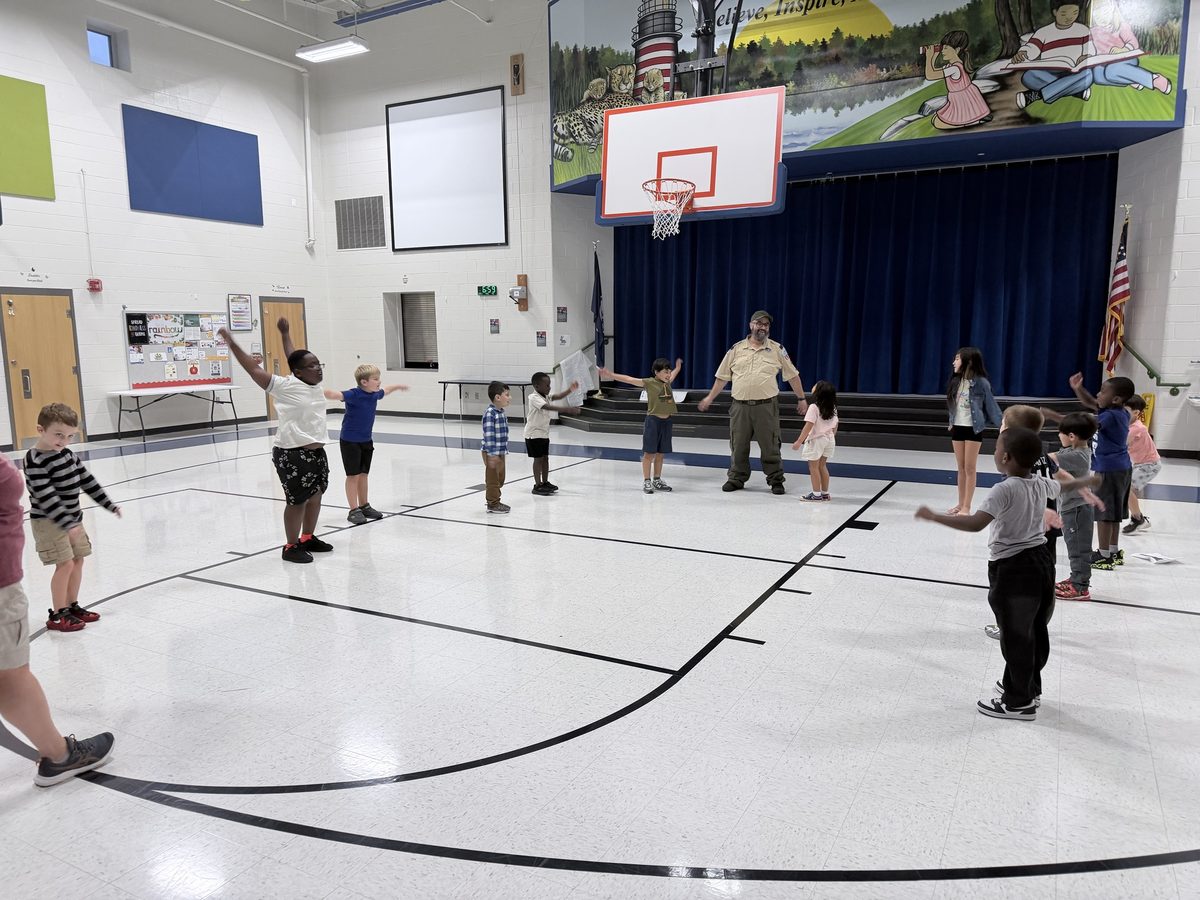 A uniformed leader leads Scouts in arm stretches in front of the gym stage, arms stretched wide.
