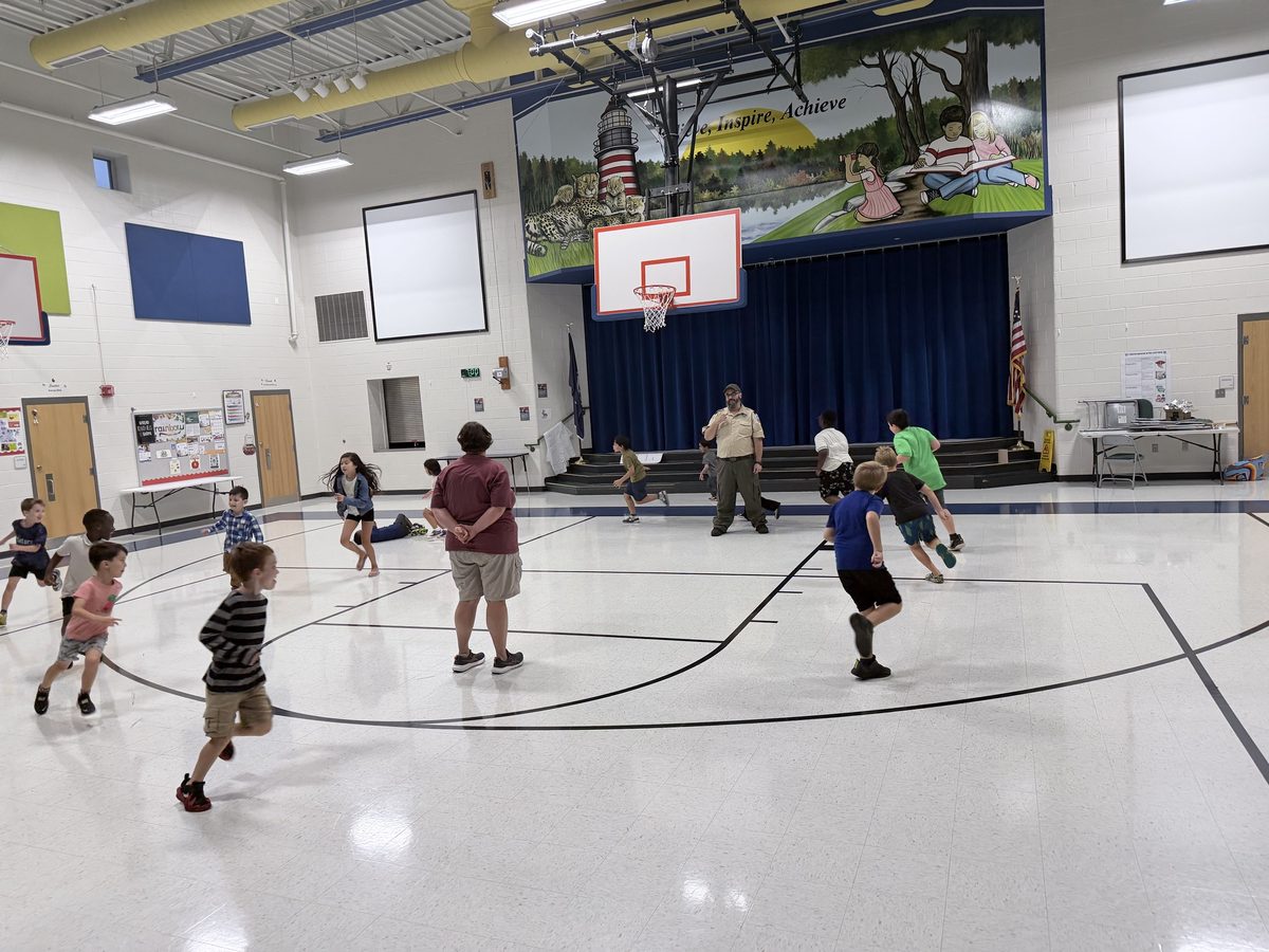 Scouts run and play a tag game across the gym while two adult leaders watch from the middle of the court.